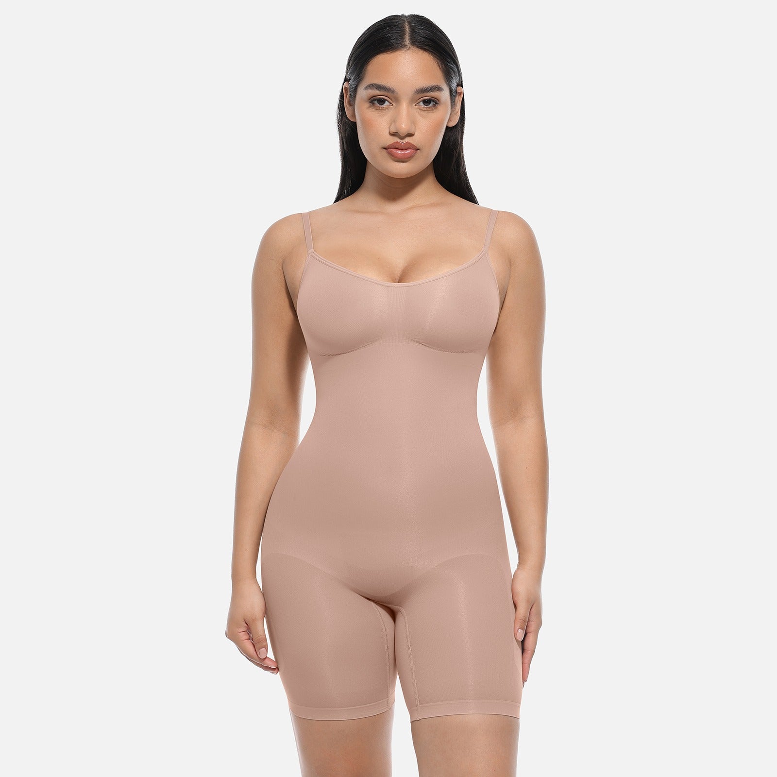 A woman modeling a nude-beige shapewear bodysuit with built-in shorts, photographed against a clean light gray studio background.