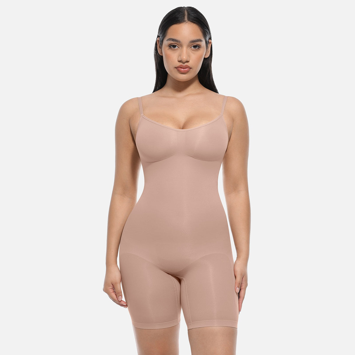 A woman modeling a nude-beige shapewear bodysuit with built-in shorts, photographed against a clean light gray studio background.